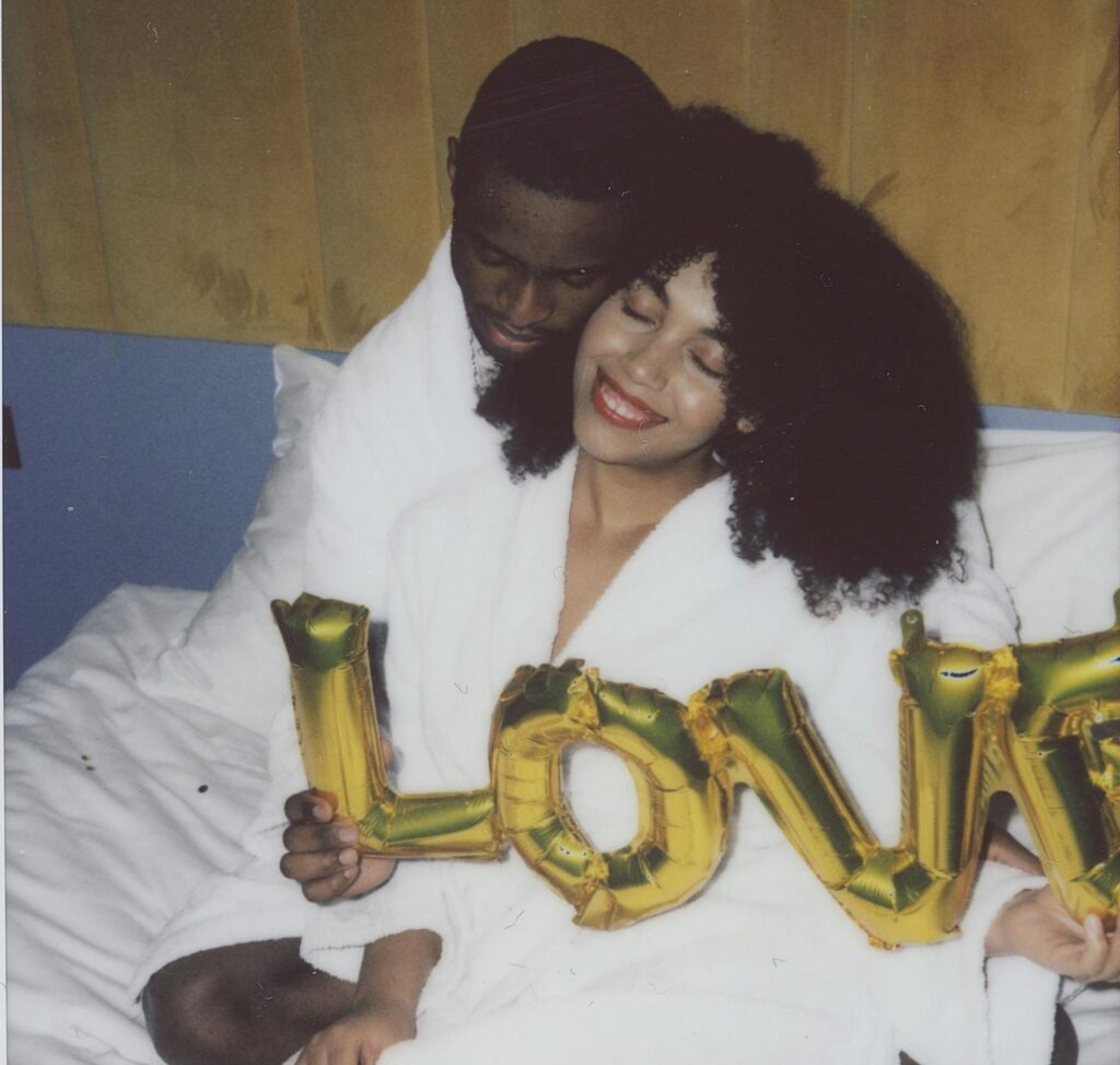 Happy couple in bathrobes holding 'LOVE' balloons, enjoying a cozy moment indoors.