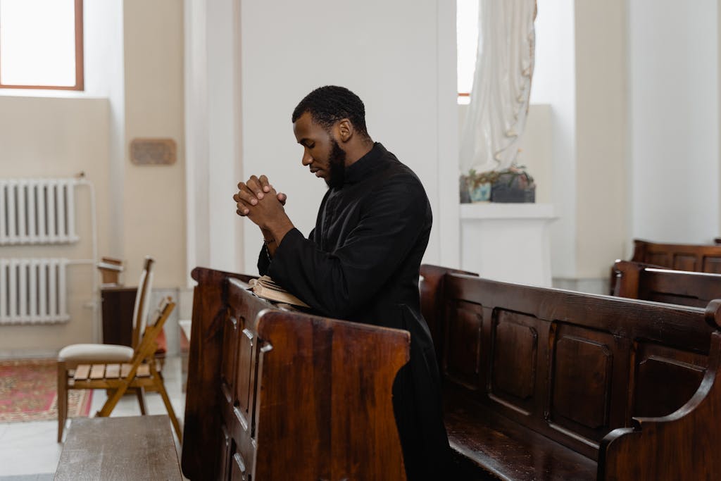 A man in a cassock kneeling and praying in a church pew, expressing deep faith.