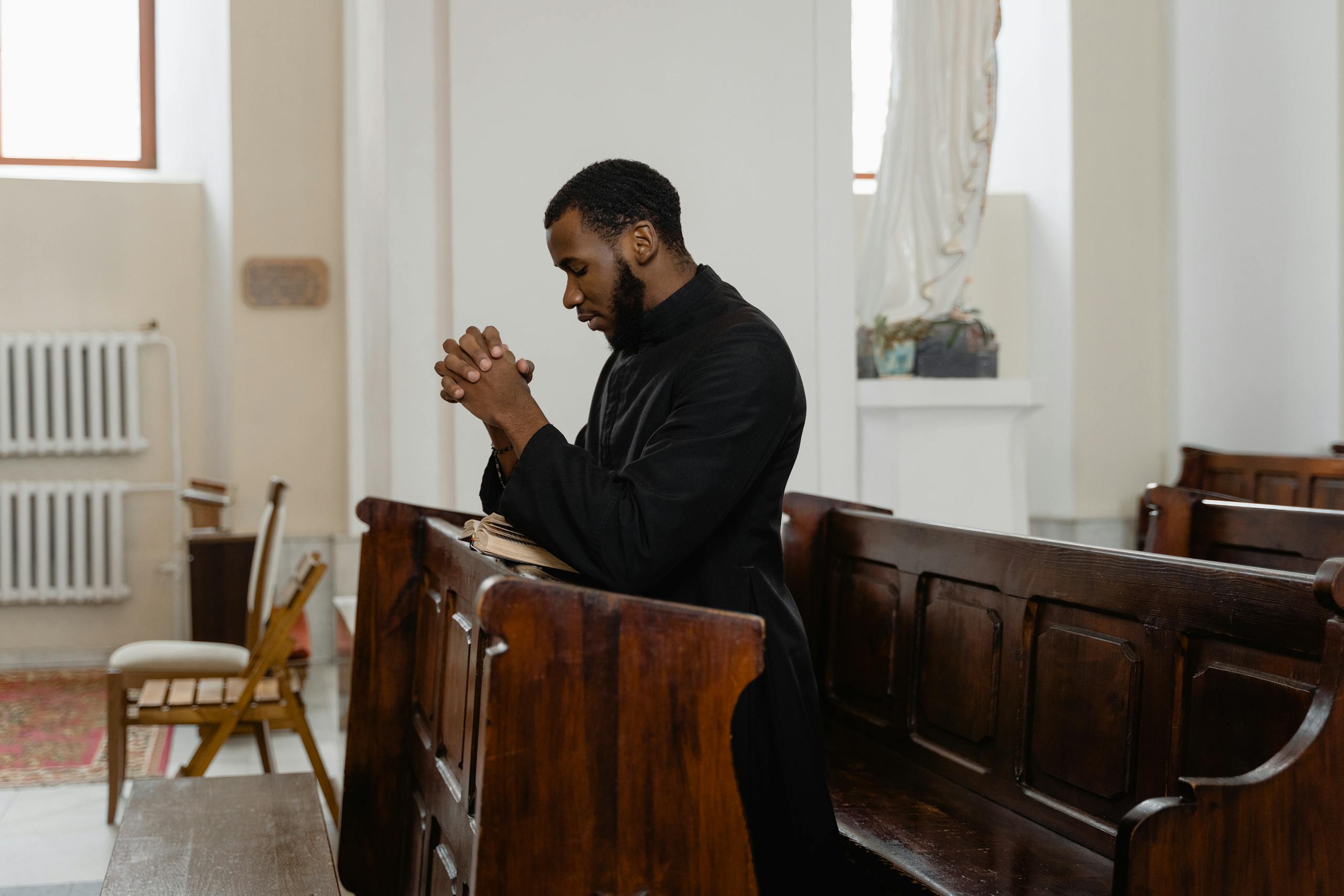 A man in a cassock kneeling and praying in a church pew, expressing deep faith.