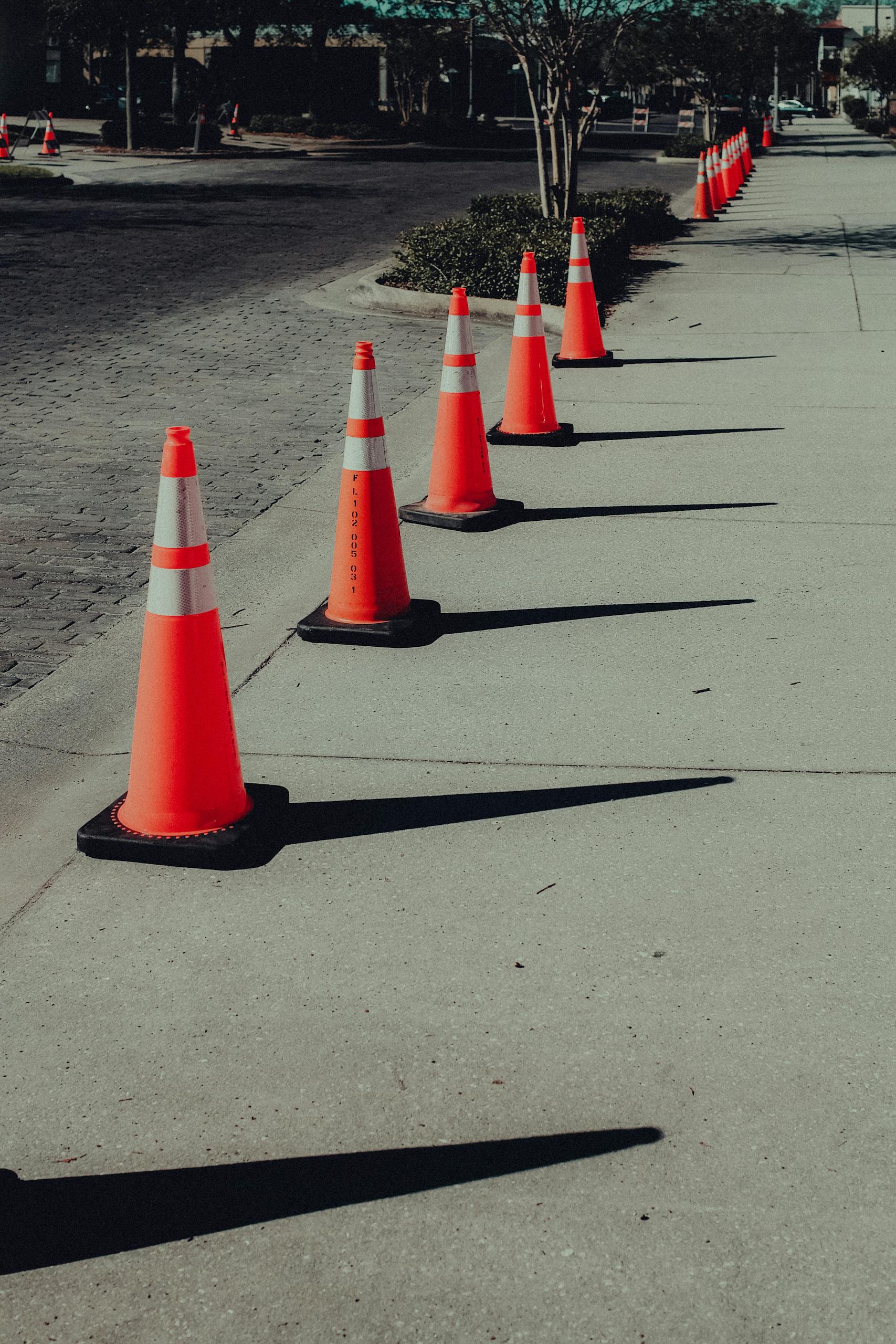 Orange traffic cones neatly arranged on a sidewalk casting shadows, in an urban setting.
