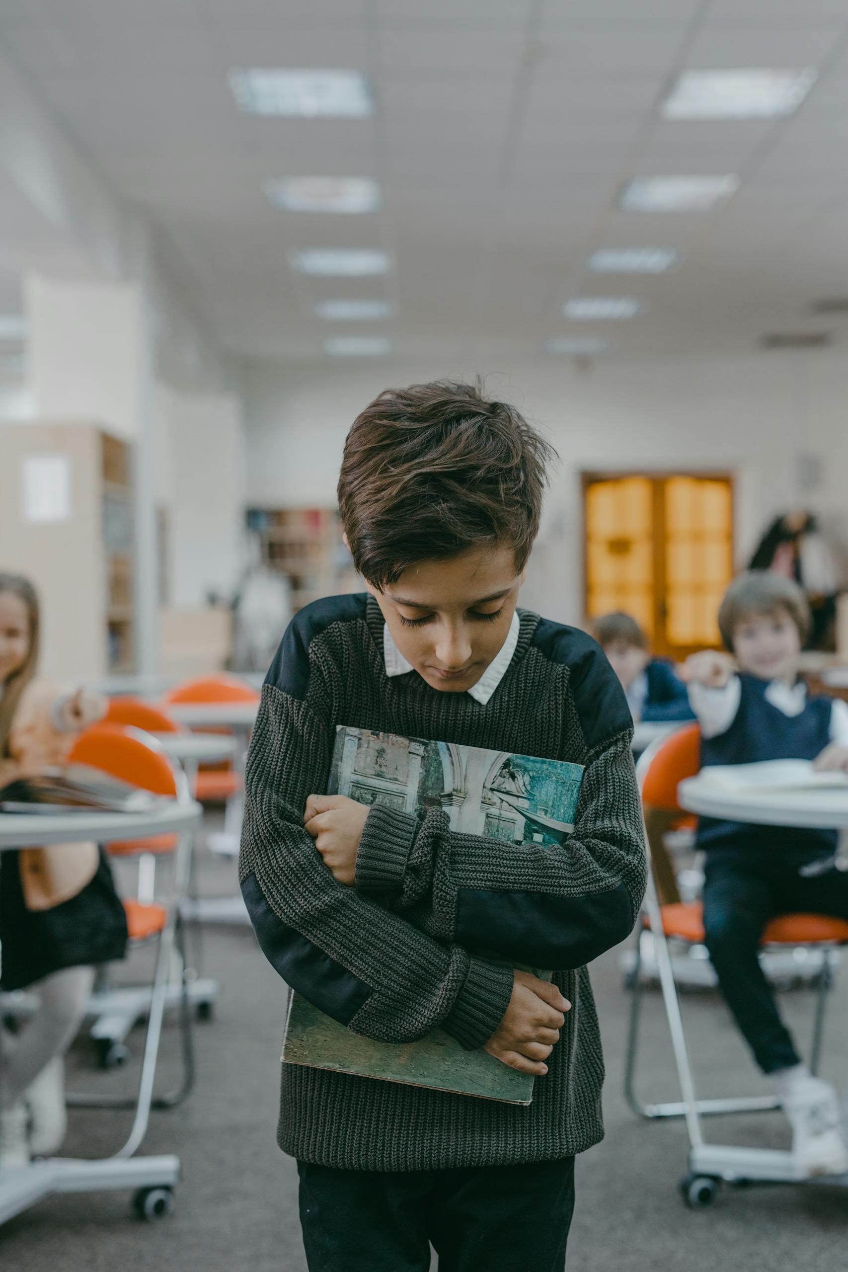 Sad boy holding books tightly, showing signs of bullying and isolation in a classroom.