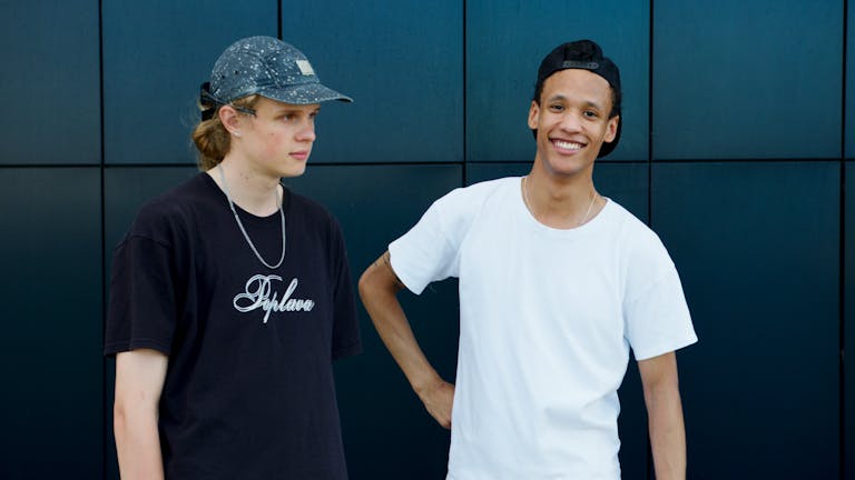 Two teenagers in casual attire, standing outdoors with a modern backdrop, smiling.