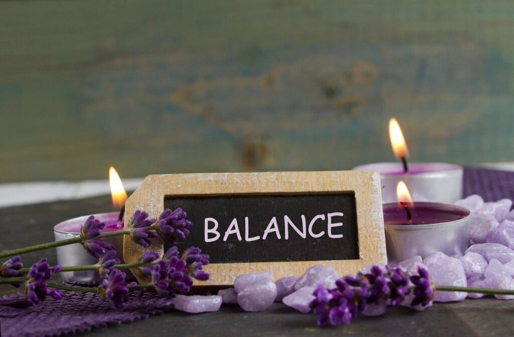 Chalkboard sign reading 'BALANCE' surrounded by purple lavender, candles, and pink-purple stones for a calming spa scene