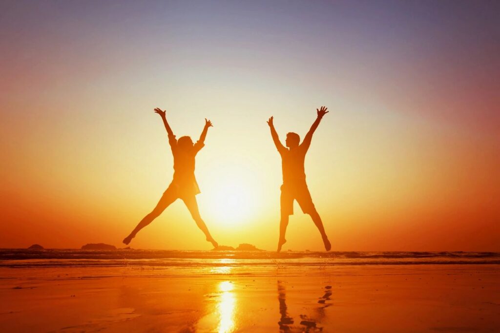Two silhouetted children jumping with arms raised on a sunset beach, reflected in the wet sand.