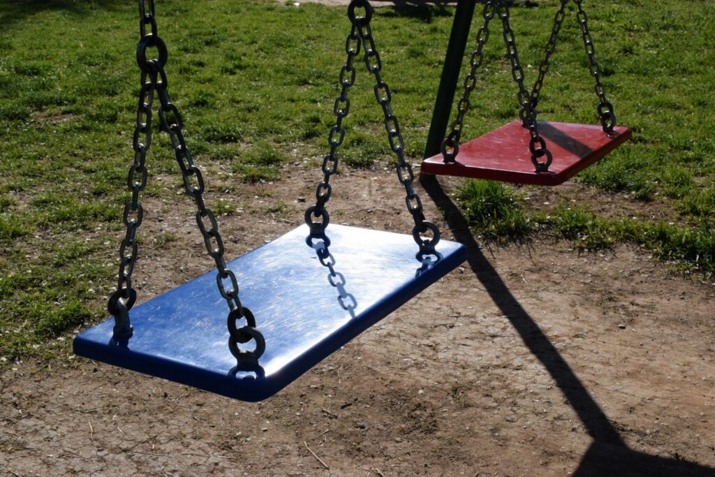 Empty blue and red swing seats hang from metal chains in a sunlit park playground.