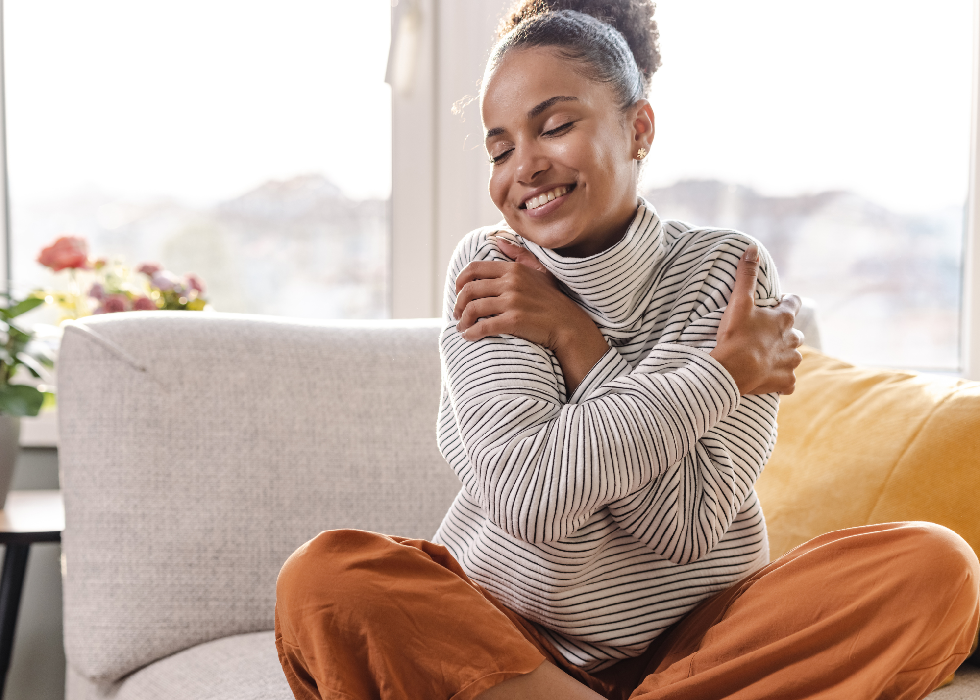 13 | Fuller Life Family Therapy Woman smiling with arms wrapped around herself while sitting on a sofa in a sunlit living room.