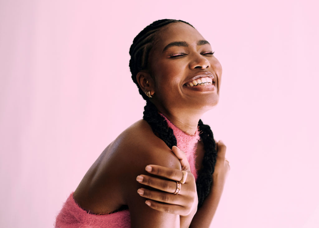 14 | Fuller Life Family Therapy Joyful close-up of a woman with braided hair and eyes closed, smiling against a pastel pink background, arms crossed over chest.