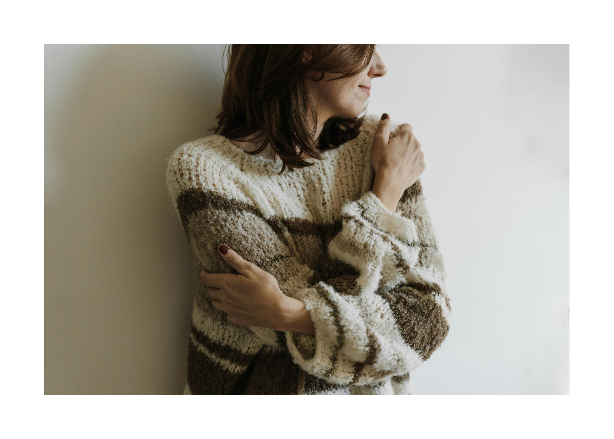 15 | Fuller Life Family Therapy Person wearing a chunky striped beige-brown sweater hugs their arms across their chest, looking to the right against a light wall.