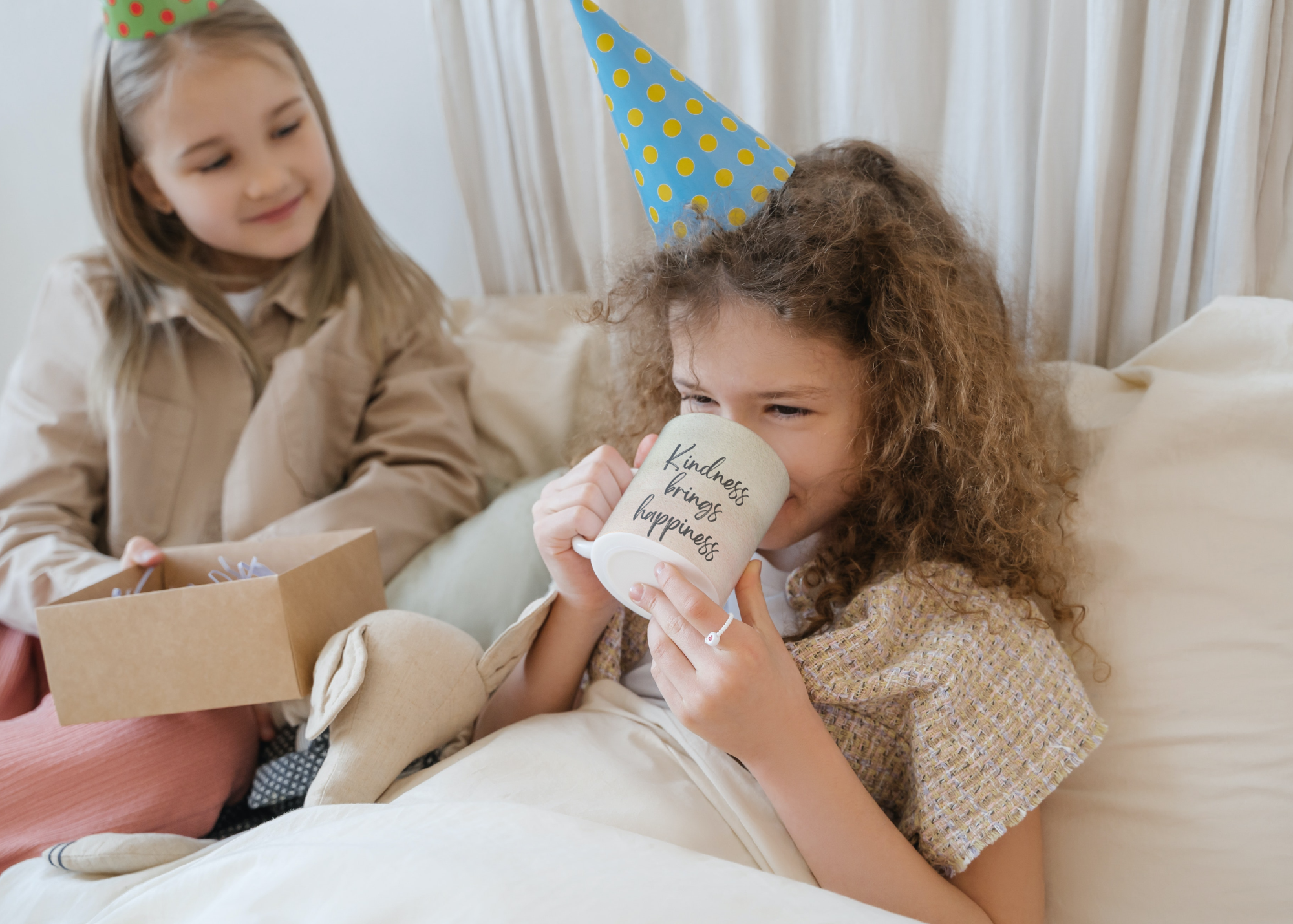 19 | Fuller Life Family Therapy Two young girls celebrate a birthday: one drinks from a mug that reads'Kindness brings happiness' while the other sits nearby with a gift bag and party hats.