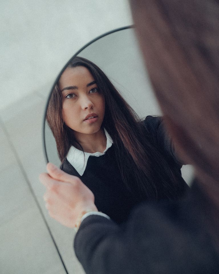 A young East Asian woman gazes into a mirror, reflecting her thoughtful expression.