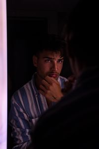Stylish young man contemplating his reflection in a dimly lit room, wearing striped shirt.
