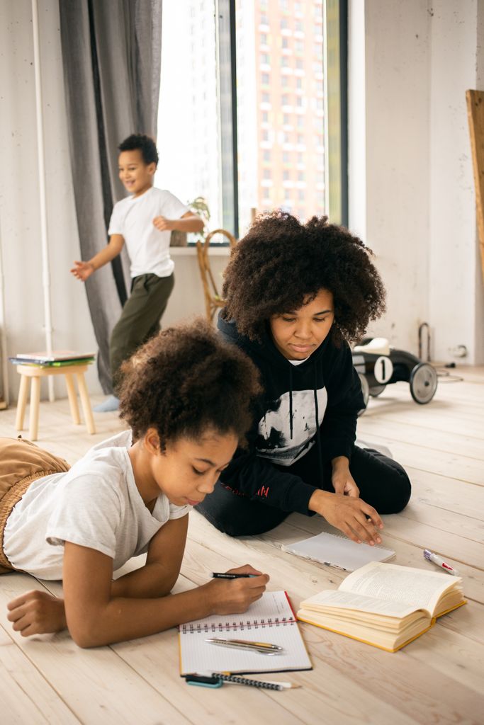 Concentrated African American mother near daughter writing in copybook while lying on wooden floor in light room with positive brother. Learned values.